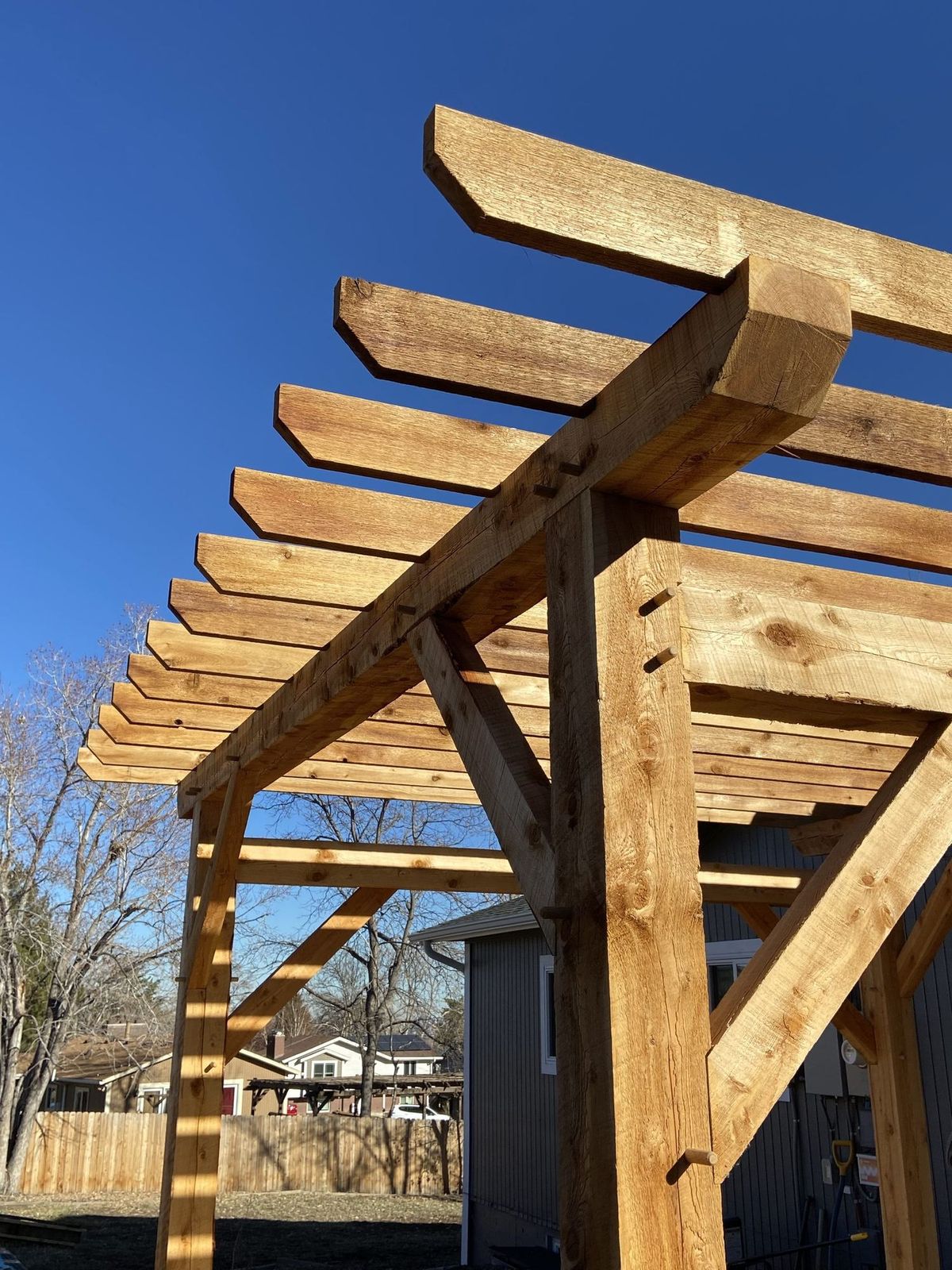 Looking straight up through the rafters of a hand-cut timber frame pergola toward a blue sky