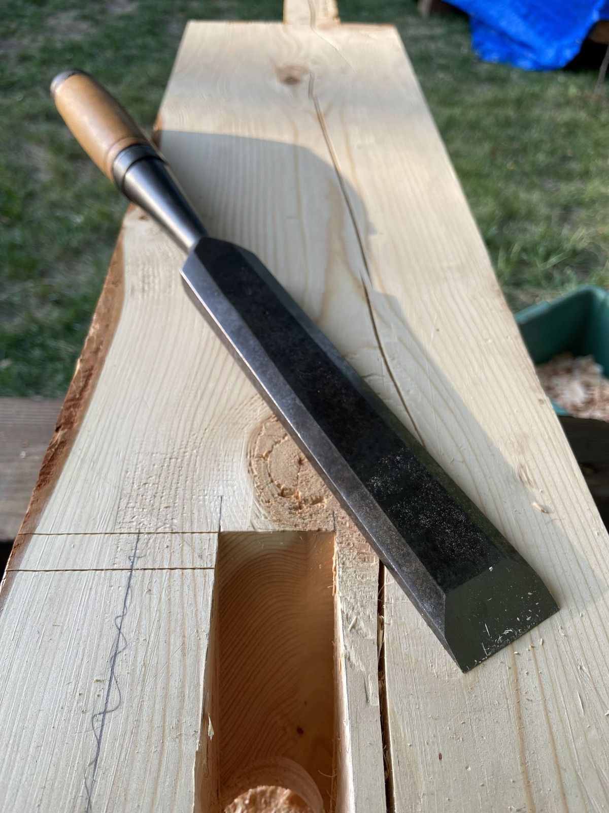 A large timber framing chisel lying across a Douglas Fir beam beside a freshly cut mortise