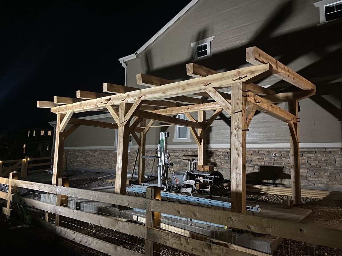Timber frame pavilion at night during installation, with green lanterns hanging from the posts