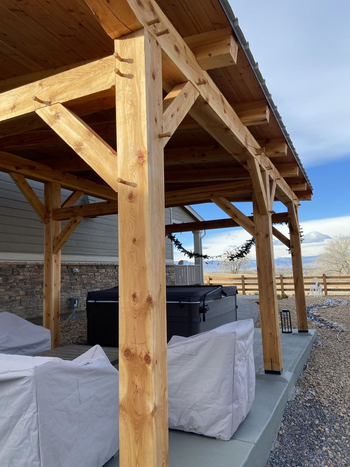 Finished timber frame pavilion beside a stone-veneer house, with a hot tub under the roof