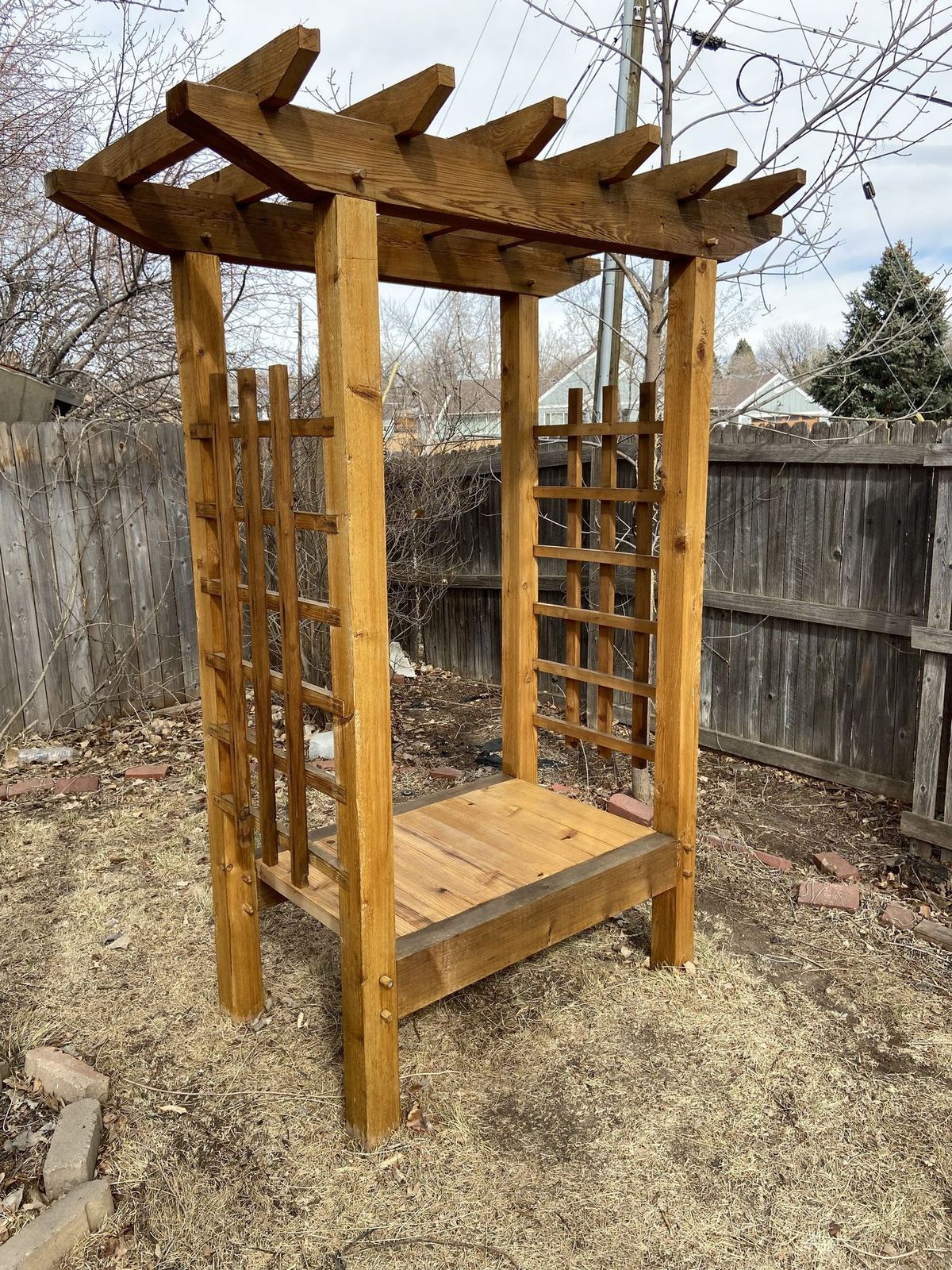 A finished cedar trellis structure with lattice panels and a plank seat in a backyard