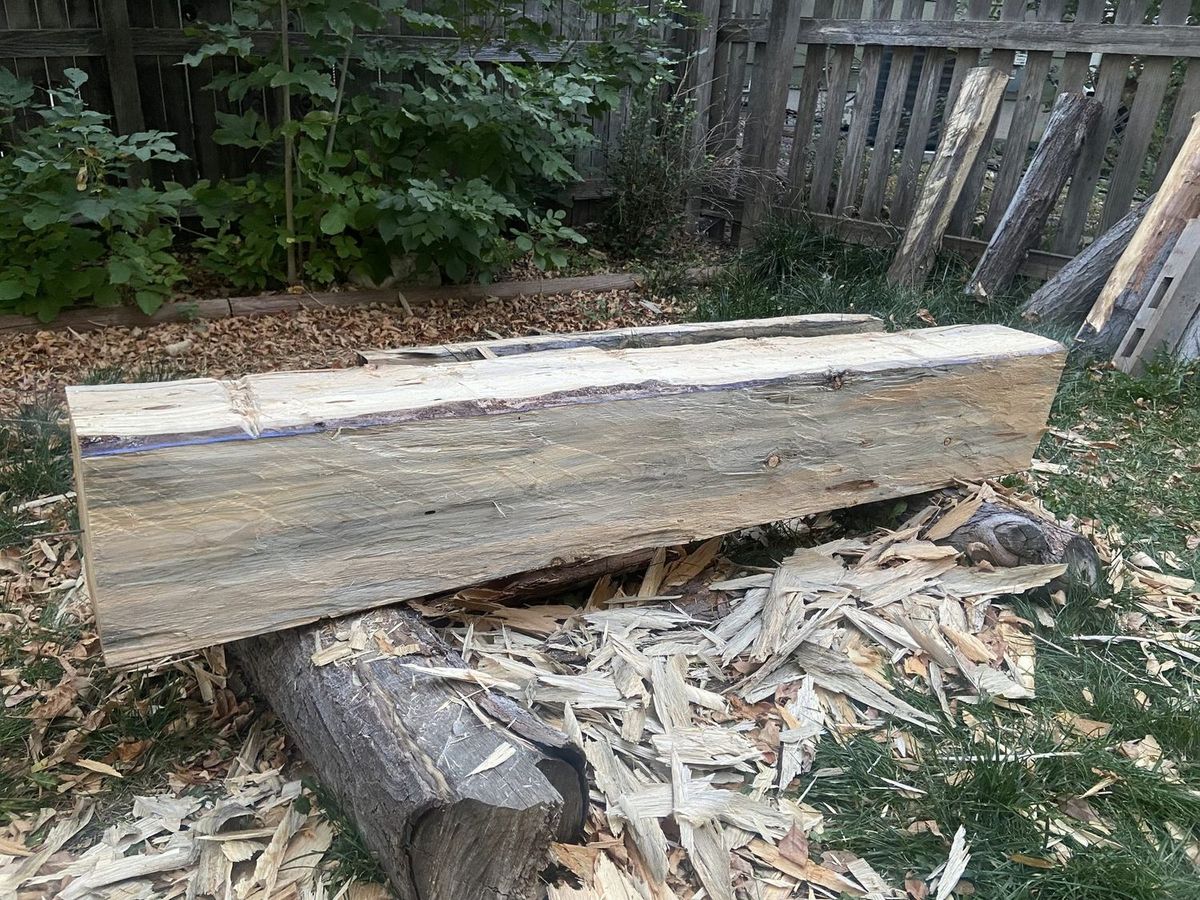 A hand-hewn timber beam with weathered gray faces lying across logs in a backyard